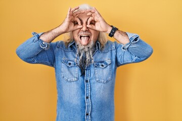 Poster - Middle age man with grey hair standing over yellow background doing ok gesture like binoculars sticking tongue out, eyes looking through fingers. crazy expression.