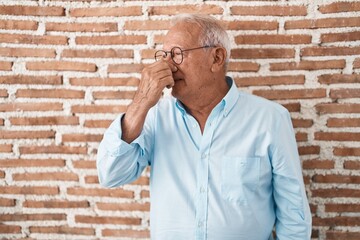 Poster - Senior man with grey hair standing over bricks wall smelling something stinky and disgusting, intolerable smell, holding breath with fingers on nose. bad smell