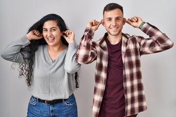 Poster - Young hispanic couple standing over white background smiling pulling ears with fingers, funny gesture. audition problem