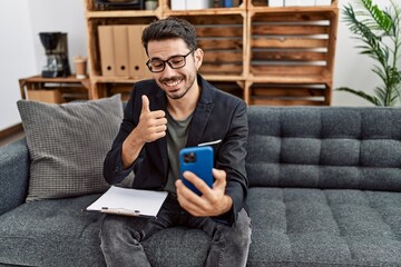 Wall Mural - Young hispanic psychologist man doing therapy on video call with smartphone smiling happy and positive, thumb up doing excellent and approval sign