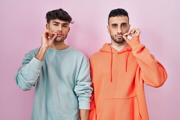 Poster - Young hispanic gay couple standing over pink background mouth and lips shut as zip with fingers. secret and silent, taboo talking