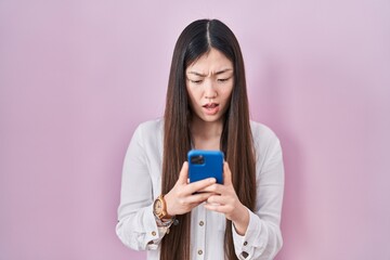 Wall Mural - Chinese young woman using smartphone typing message in shock face, looking skeptical and sarcastic, surprised with open mouth