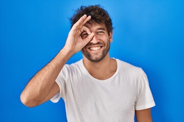 Poster - Hispanic young man standing over blue background doing ok gesture with hand smiling, eye looking through fingers with happy face.