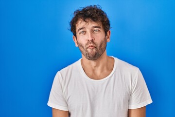 Wall Mural - Hispanic young man standing over blue background making fish face with lips, crazy and comical gesture. funny expression.