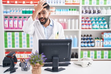 Wall Mural - Hispanic man with beard working at pharmacy drugstore surprised with hand on head for mistake, remember error. forgot, bad memory concept.