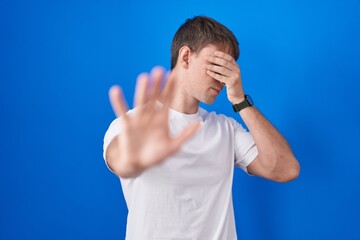 Poster - Caucasian blond man standing over blue background covering eyes with hands and doing stop gesture with sad and fear expression. embarrassed and negative concept.