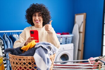 Sticker - Young brunette woman with curly hair doing laundry using smartphone smiling happy and positive, thumb up doing excellent and approval sign