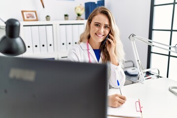 Wall Mural - Young woman wearing doctor uniform writing on document talking on smartphone at clinic