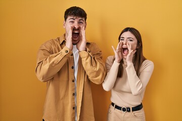 Sticker - Young hispanic couple standing over yellow background shouting angry out loud with hands over mouth