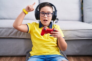 Poster - Young hispanic kid playing video game holding controller wearing headphones smiling amazed and surprised and pointing up with fingers and raised arms.