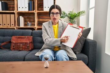 Poster - Young hispanic psychiatrist woman working at consultation office with clipboard in shock face, looking skeptical and sarcastic, surprised with open mouth