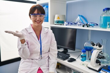 Canvas Print - Young brunette woman working at scientist laboratory smiling cheerful presenting and pointing with palm of hand looking at the camera.