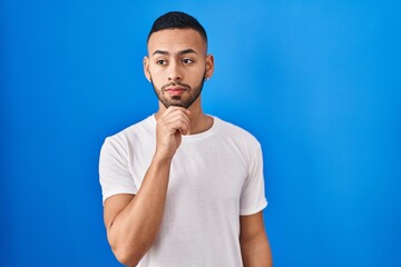 Canvas Print - Young hispanic man standing over blue background thinking concentrated about doubt with finger on chin and looking up wondering