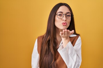 Poster - Young brunette woman standing over yellow background wearing glasses looking at the camera blowing a kiss with hand on air being lovely and sexy. love expression.