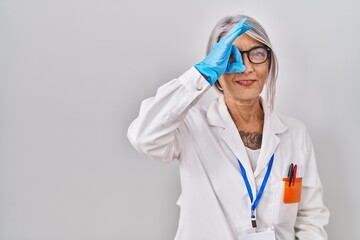 Wall Mural - Middle age woman with grey hair wearing scientist robe doing ok gesture with hand smiling, eye looking through fingers with happy face.