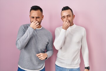 Poster - Homosexual couple standing over pink background smelling something stinky and disgusting, intolerable smell, holding breath with fingers on nose. bad smell