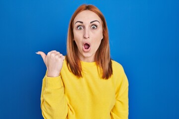 Wall Mural - Young woman standing over blue background surprised pointing with hand finger to the side, open mouth amazed expression.