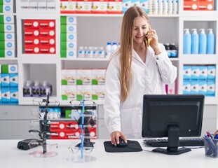 Wall Mural - Young caucasian woman pharmacist talking on smartphone using computer at pharmacy