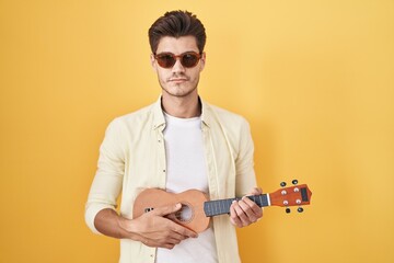 Canvas Print - Young hispanic man playing ukulele over yellow background relaxed with serious expression on face. simple and natural looking at the camera.