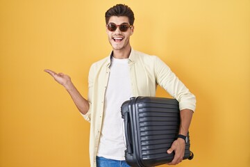 Wall Mural - Young hispanic man holding suitcase going on summer vacation pointing aside with hands open palms showing copy space, presenting advertisement smiling excited happy