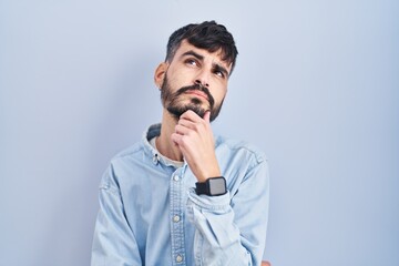 Wall Mural - Young hispanic man with beard standing over blue background with hand on chin thinking about question, pensive expression. smiling with thoughtful face. doubt concept.