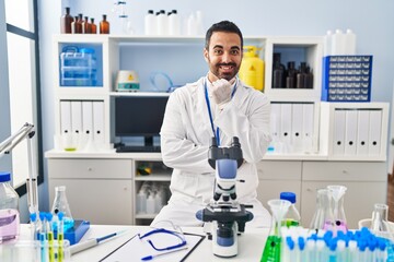 Canvas Print - Young hispanic man with beard working at scientist laboratory looking confident at the camera smiling with crossed arms and hand raised on chin. thinking positive.