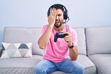 Canvas Print - Hispanic young man playing video game holding controller sitting on the sofa yawning tired covering half face, eye and mouth with hand. face hurts in pain.