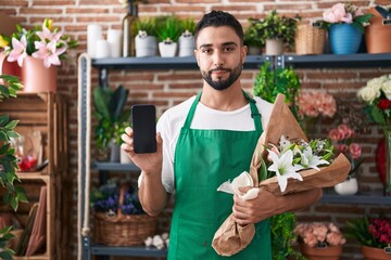 Wall Mural - Hispanic young man working at florist shop showing smartphone screen relaxed with serious expression on face. simple and natural looking at the camera.