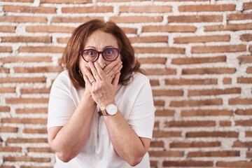 Sticker - Senior woman with glasses standing over bricks wall shocked covering mouth with hands for mistake. secret concept.