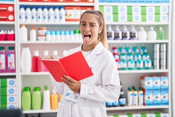 Poster - Young blonde woman working at pharmacy drugstore holding notebook sticking tongue out happy with funny expression.