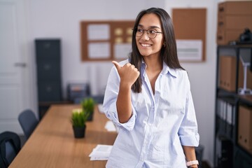 Canvas Print - Young hispanic woman at the office smiling with happy face looking and pointing to the side with thumb up.