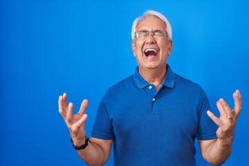 Sticker - Middle age man with grey hair standing over blue background celebrating mad and crazy for success with arms raised and closed eyes screaming excited. winner concept