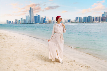 Wall Mural - a young indian woman walks along the beach, taking in the breathtaking sea view of Abu Dhabi's towering skyscrapers.