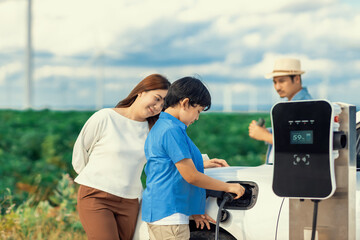 Concept of progressive happy family enjoying their time at wind farm with electric vehicle. Electric vehicle driven by clean renewable energy from wind turbine generator for charging station.