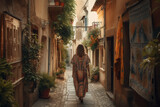 Fototapeta Uliczki - Boho girl walking at a quaint village with narrow streets and charming storefronts, rear view. Travel light with ease and let nature guide you concept. Generative AI