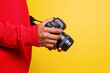 Side view man photographer in red shirt makes photos isolated on yellow background. Male hands hold the camera and adjust lens. Digital single-lens reflex camera in hands.