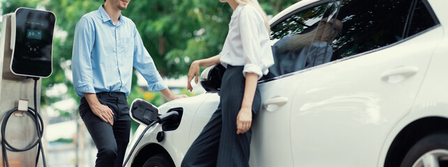 Progressive businessman and businesswoman with electric car parking and connected to public charging station before driving around city center. Eco friendly rechargeable car powered by clean energy.