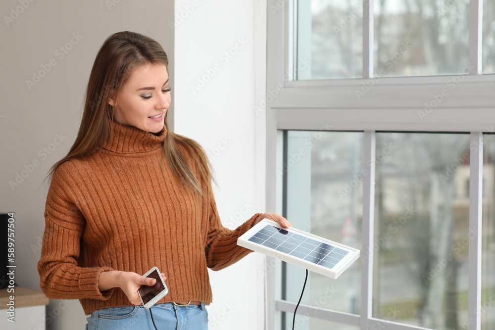 Pretty young woman charging mobile phone with portable solar panel near window