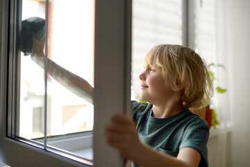 Cute little boy washing a window at home. Child helping parents with household chores, for example, cleaning windows in his house. Children doing housework.