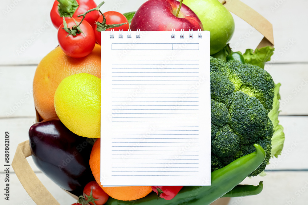 Paper bag with vegetables, fruits and blank notebook on white wooden background