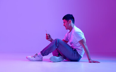 Happy, smiling young man in casual clothes sitting on floor, looking on mobile phone, texting messages against purple background in neon light. Concept of human emotions, youth, fashion, lifestyle