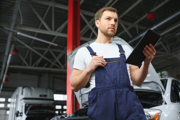 Bearded mechanic in overalls standing in garage of a car salon and holding tablet. He is about to diagnostic breakdown.