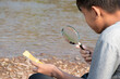Asian boy sits holding a magnifying glass and looking at a convex lens to try burning a small piece of paper from the sun's rays near a river in the afternoon during school holidays to do experiment.