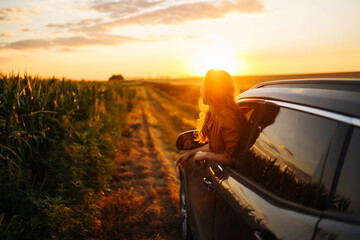 Wall Mural - Young happy woman leaning out of the car window enjoying the sunset. The concept of active lifestyle, travel, tourism, nature.