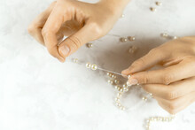 Close-up Of Woman Hands Stringing Pearls On A Necklace On White Background. Beads Jewellery Making Process. Hobby Handmade Concept. Top Down View. Selective Focus. Blurred Background.