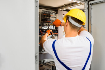 Electrician with screwdriver tighten up switching electric actuator equipment in fuse box. View from the back, a young specialist in a yellow hard hat and overalls. Indoor.