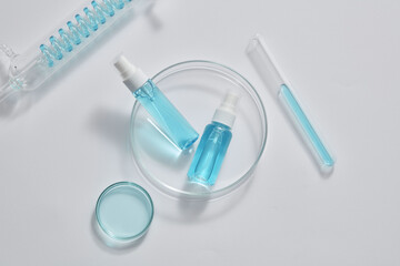 Top view of unlabeled mist spray bottles placed on a transparent dish. A spiral pipe tube, petri dish and a test tube of blue fluid are arranged. Mockup of skin care product