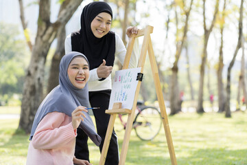 Two Asian young muslim women wear hijab drawing water color on white paper on easel outdoor in garden. Group of Asian muslim women learning painting art lesson outside classroom with happy and smiling