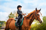 Small child in jockey outfit is riding horse on blue sky with clouds background. School of riding and equestrian sports.