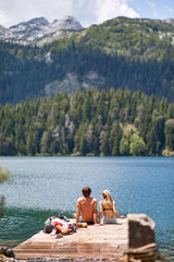 Wall Mural - Young couple sitting on wooden jetty by lake. Summer trip in nature. Lifestyle, love, togetherness, nature concept